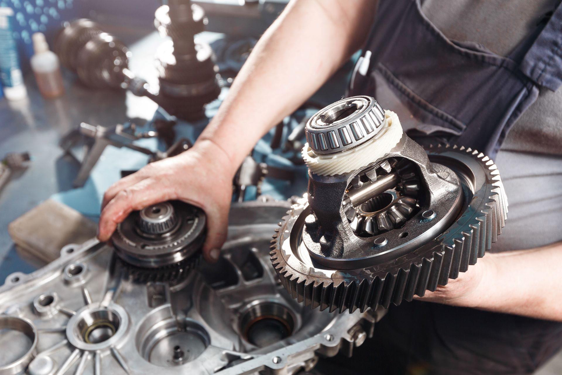 Ein Mechaniker hält ein zerlegtes Getriebe in der Hand, das aus Metallzahnrädern und Lagern besteht. Im Hintergrund sind Werkzeuge und weitere Getriebekomponenten zu sehen. Das Bild zeigt die komplexe Technik und Präzision der Antriebstechnik