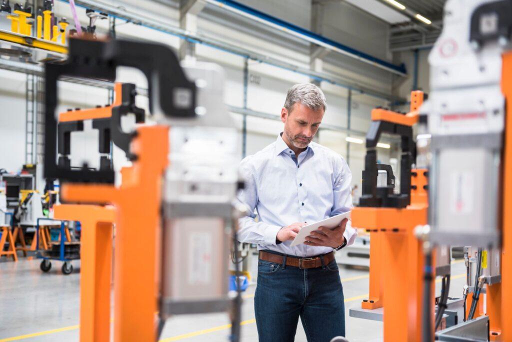 Ingenieur mit Tablet zwischen Maschinen in Produktionshalle analysiert Vorrichtungsbau fuer Fertigungsprozesse